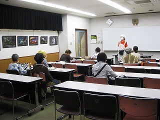 複数の受講者が教室の机に座り、前方で講師がホワイトボードの前に立って講義を行っている様子を捉えた写真