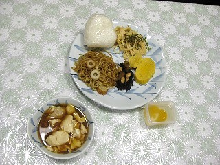 柄入りのテーブルクロスが敷かれたテーブルに、おにぎりや焼きそば、スパゲッティなどの料理が盛り合わせられたプレートと、煮物、デザートが並べられた様子を撮影した写真