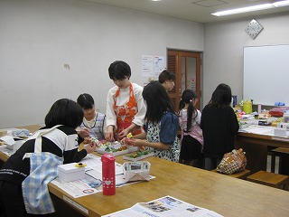 教室で数名の生徒が机を囲んで座り、一人の女性が中央で熱心に生徒たちに手芸を教えている様子を捉えた写真