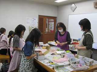 教室内で複数の女性と子供たちが机を囲んで座り、手芸の作業を熱心に行っている様子を撮影した写真