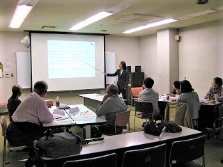 室内で、プロジェクターに映し出されたスライドをスーツ姿の女性が指し示して説明しており、複数の参加者が熱心に話を聞いている様子の写真