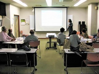 室内で、講師がスクリーンの前で説明をしており、それに対し数名の参加者が着席して耳を傾けている様子の写真