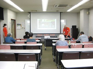 災害関連死についてプロジェクターで投影されたスライドを講師が説明しており、複数の参加者がテーブルについて真剣な表情で話を聞いている様子の写真