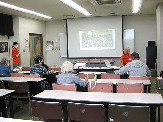 学校の教室のような室内で、講師と思われる男性がプロジェクターの前に座り、災害関連死について説明しており、複数の参加者が席について熱心に話を聞いている様子の写真