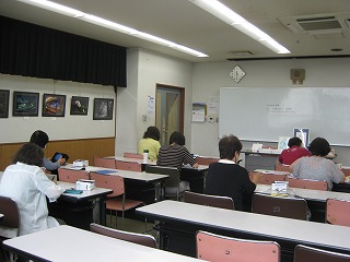 複数の参加者が前方のホワイトボードに向かって座っており、両側の壁には写真や絵画が飾られている、講座の様子の写真