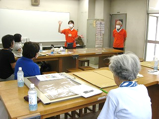 複数の参加者が机に座り、赤いポロシャツの女性がホワイトボードの前で手を上げて説明しており、その隣に男性が立っている、イベント会場の様子の写真