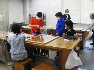 白い部屋で複数の大人がテーブルを囲んでおり、赤いポロシャツの男性が何かの作品をビニールで梱包し、他の参加者がその様子を見ている様子の写真