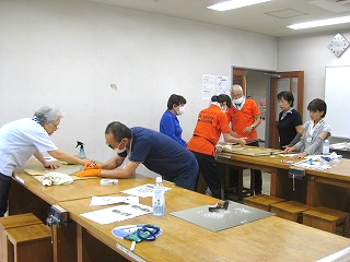 複数の大人たちがテーブルを囲んで何かを制作しており、赤いポロシャツの男性が他の参加者に手伝いながら指導している様子の写真