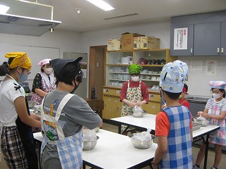 料理教室の調理台を囲んで、三角巾とエプロンをつけた複数人の親子が、ボウルを使って調理をしながら、講師が子供たちに作り方を説明している様子の写真