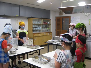 学校の家庭科室のような室内で、三角巾とエプロンをつけた複数人の子供たちが、ボウルやビニール袋を使って、講師に教えてもらいながら調理している様子の写真