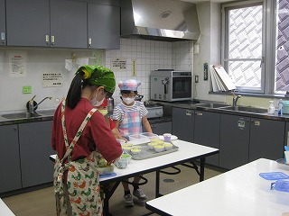 厨房内で三角巾とエプロンをつけた2人の子供がテーブルに向かい、1人の子供が何かを混ぜており、もう1人が完成したプリンのようなお菓子を並べている様子の写真