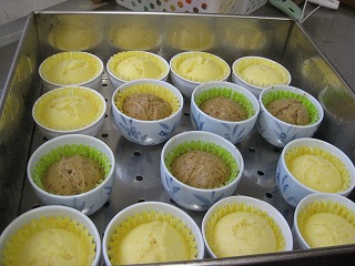 せいろの中に黄色と茶色の蒸しパンが、それぞれカップに入れられて並んでおり、ふっくらと膨らんだ出来立ての様子を上から撮影した写真