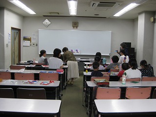 学校のような教室で、複数の子供たちが机に向かい、先生や保護者と思われる大人が、ホワイトボードの前で子供たちに熱心に指導している様子の写真