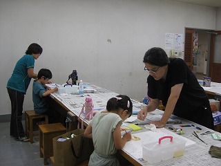 新聞紙が敷かれた長いテーブルで、大人2人と子供2人がそれぞれ座って絵を描いたり工作をしたりしており、大人が子供にやり方を教えている様子の写真