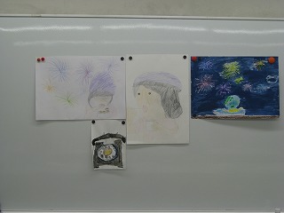 白い壁に、花火と人物のイラストが複数枚、素朴なタッチで描かれた子供たちの作が画鋲で貼り付けられている写真