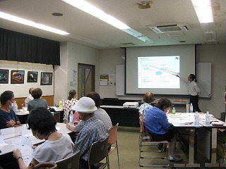 室内の会議室で、大勢の高齢者が2つのテーブルに分かれて座り、プロジェクターで投影されたスライドを女性講師が指し棒を使って説明している様子の写真