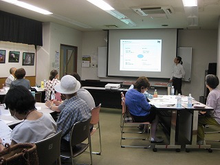 広めの室内で複数の参加者がテーブルに分かれて座り、プロジェクターで投影された画面を女性講師が指し示して説明している様子の写真