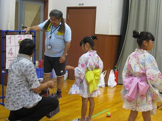 室内のイベントで、浴衣を着た2人の女の子が大人と一緒にレクリエーションに参加しており、大人の男性がボードを指差しながら説明している様子の写真