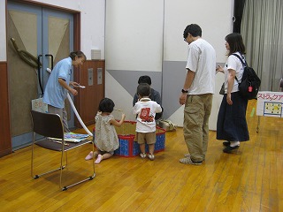 イベント会場の室内で、複数の親子が子供向けのレクリエーションコーナーに集まり、子供たちが遊ぶ様子を大人が見守りながら話している様子の写真