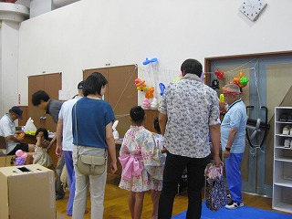 風船で飾られたイベント会場で、浴衣を着た女の子の後ろ姿を中心に、複数の親子連れが集まって楽しんでいる様子の写真