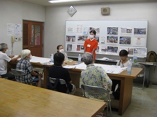 室内でテーブルを囲んだ人々が高齢者向けの説明会に参加し、女性の講師がマスクを着用してホワイトボードの前で話している様子の写真