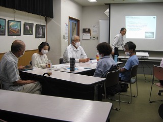 教室で、プロジェクターで資料が投影されたスクリーンの横で講師が話しているのをマスクを着用した数人の参加者がテーブルを囲んで座り話を聞いている写真