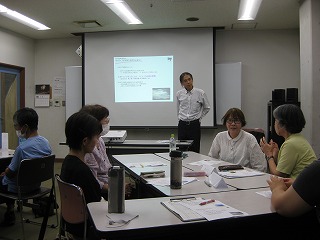 教室で、プロジェクターに資料を投影しながら立った姿勢で説明している男性講師と、テーブルを囲んで熱心に話し合ったり耳を傾けたりしている参加者の写真