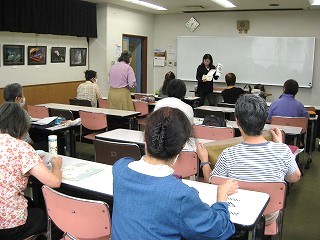 教育施設のような教室で、マスクを着用した参加者らが机に座り、教壇に立つ女性の教師が白いホワイトボードの前で教科書のようなものを手に持ち授業を行っている写真