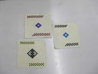 白い布地に赤茶色、緑色、黒色、青色などのチェック柄と菱形の模様の「こぎん刺し」された正方形のコースターが3枚写っている写真