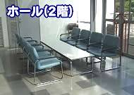 光沢のある床の上に、シルバーのフレームと青いビニールレザーの椅子が長机を挟んで向かい合わせに複数並んでおり、左上に「ホール(2階)」の文字が重ねられた施設の休憩スペースの写真