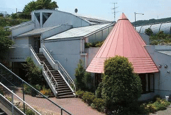 白い建物に屋外階段があり、右側に赤い円錐形の屋根を持つ建物が併設された、周囲に緑の植栽が配置された鶴巻公民館の外観写真