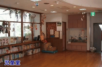大きな窓の前に低い本棚が並び、その上に装飾が施され、右奥には茶色の収納棚とカウンターがある、天井の照明と木製の床が特徴的な「児童室」と書かれた室内の写真