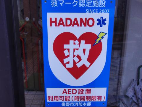 2007年から救マーク認定施設であることと、秦野市のロゴデザインが入り、中央の赤いハートの中に「救」の白い文字があり、AED設置と利用可能時間制限を明記した看板の写真