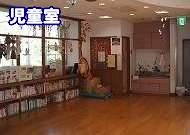 フローリングの床と大きな窓を持つ明るい部屋で、窓際に背の低い本棚が並び、遊具や装飾品が見え、部屋の左上には「児童室」という文字が重ねられた施設の写真