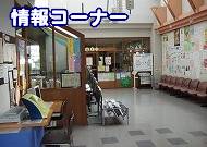 広い施設内のホールの一角を捉えた写真で、手前に木製の机や椅子、掲示板、情報誌のラックなどが設置され、壁にはポスターが貼られ、左上には「情報コーナー」の文字が重ねられている屋内写真