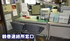 透明なアクリル板が設置されたカウンター越しに、緑色のキャビネット、コンピューターモニター、電話機、複数の書類や掲示物が見え、左下に「税務連絡所窓口」という文字が入った鶴巻連絡所窓口の写真