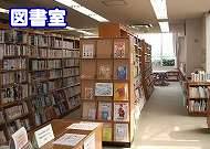 木製の背の高い書棚が両側に並び、奥の窓から光が差し込む通路があり、手前の書棚の一部には表紙を前に向けた展示棚があり、左上には「図書室」という文字が重ねられた図書室内の写真
