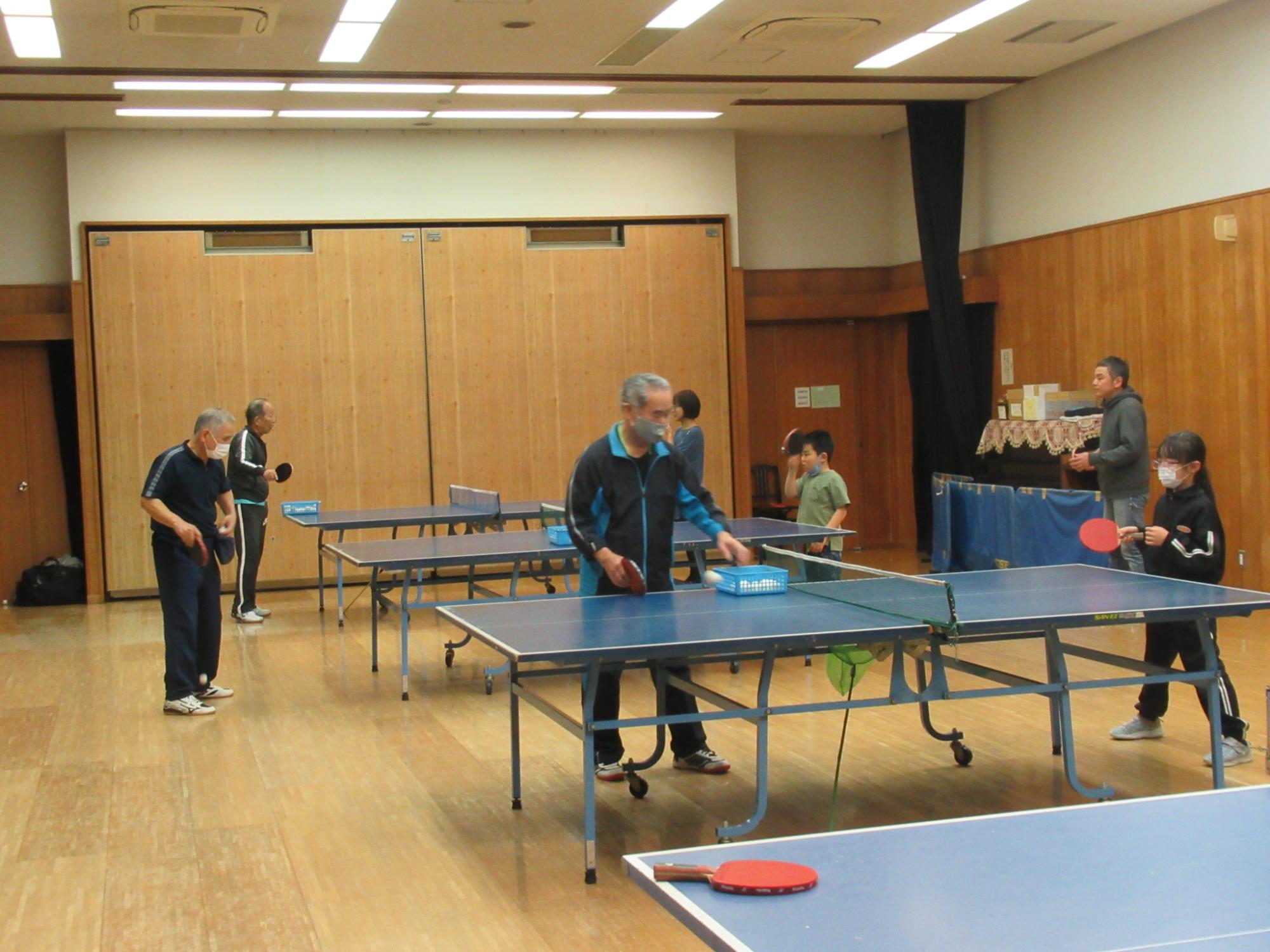 木製の壁面と白い天井を持つ室内に複数の卓球台が設置され、マスクを着用した参加者が卓球を楽しんでいる様子の写真