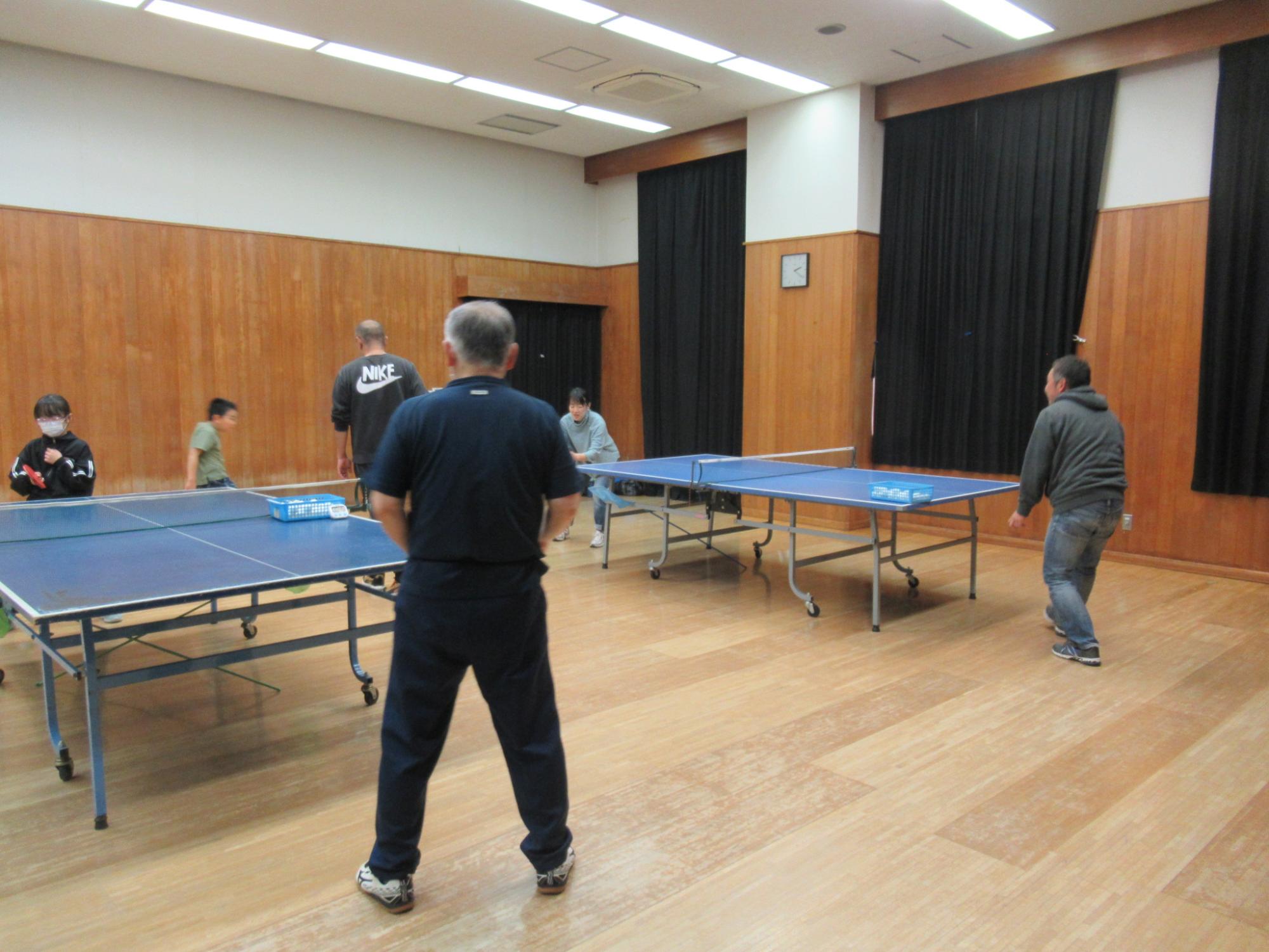 木製床の体育館内に複数の青い卓球台が設置され、木目調の壁面と黒いカーテン、白い天井照明のある室内で複数の参加者が卓球を楽しんでいる写真
