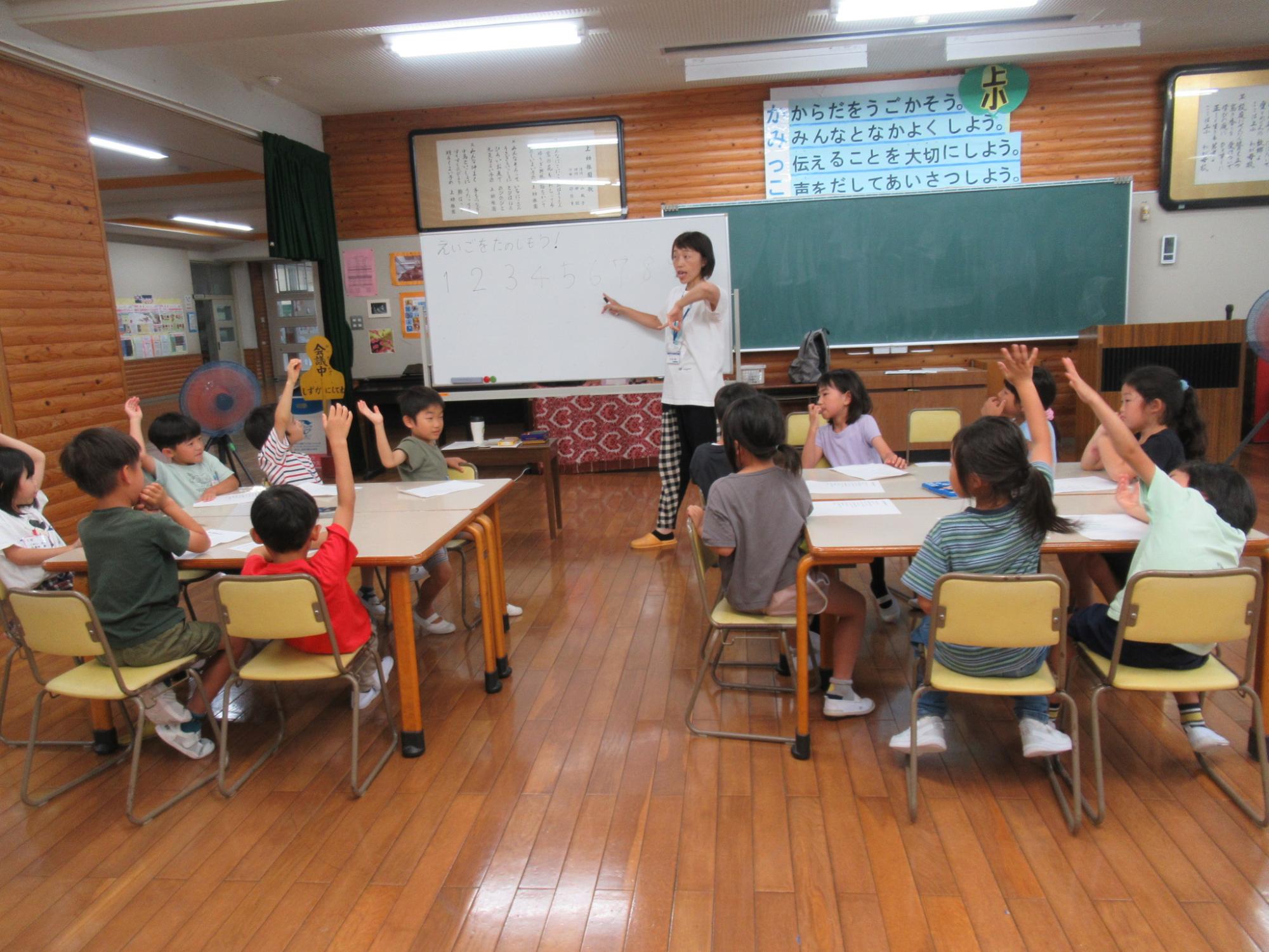 木目調の壁の教室で、女性教師がホワイトボードに数字を書きながら指導し、その周りの小さな椅子と机に座った10人以上の小学生ほどの子どもたちが、一斉に積極的に手を挙げて発言しようとしている授業風景の写真