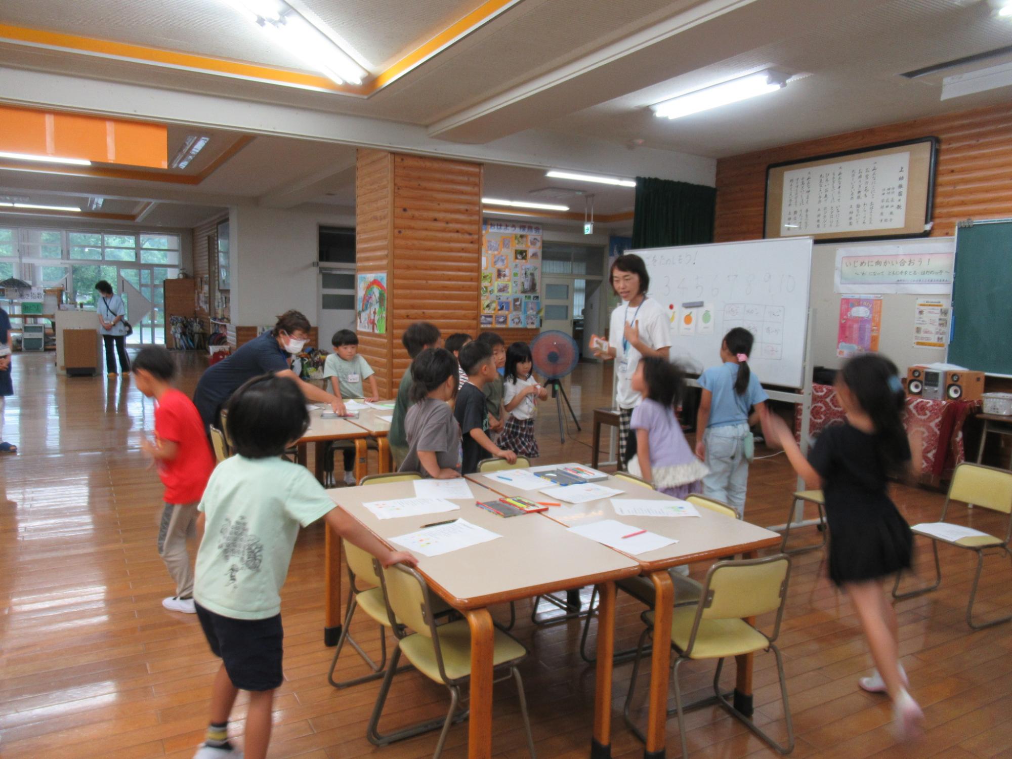 木目調の壁と明るい蛍光灯のある室内で、女性教師がホワイトボードの前で指導し、机の上には画用紙のような紙が広げられ、小学校低学年と思われる複数の児童が活発に動き回ったり、教師の話を聞いたりしている賑やかな学習風景の写真
