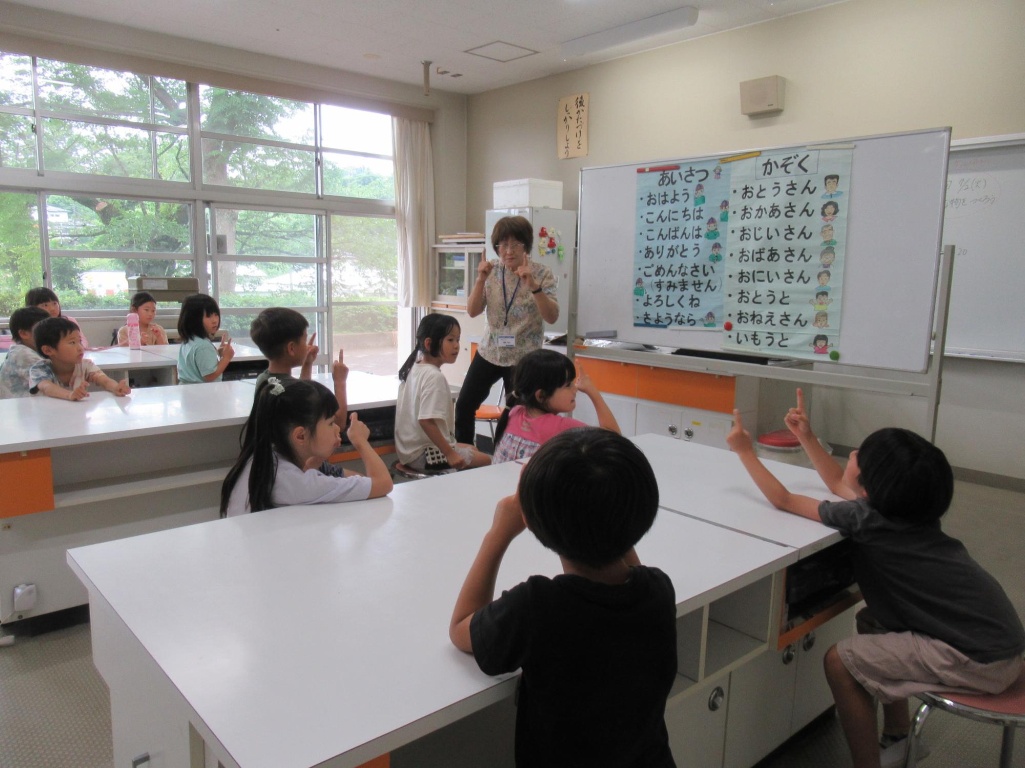学校の教室のような室内で、窓際に並べられた白い机の前に座る9人の小学生ほどの子どもたちが、挨拶や家族の名前などの日本語の単語が表示されたホワイトボードの前で、身振り手振りを交えて指導する女性教師の授業を受けている風景を写した写真
