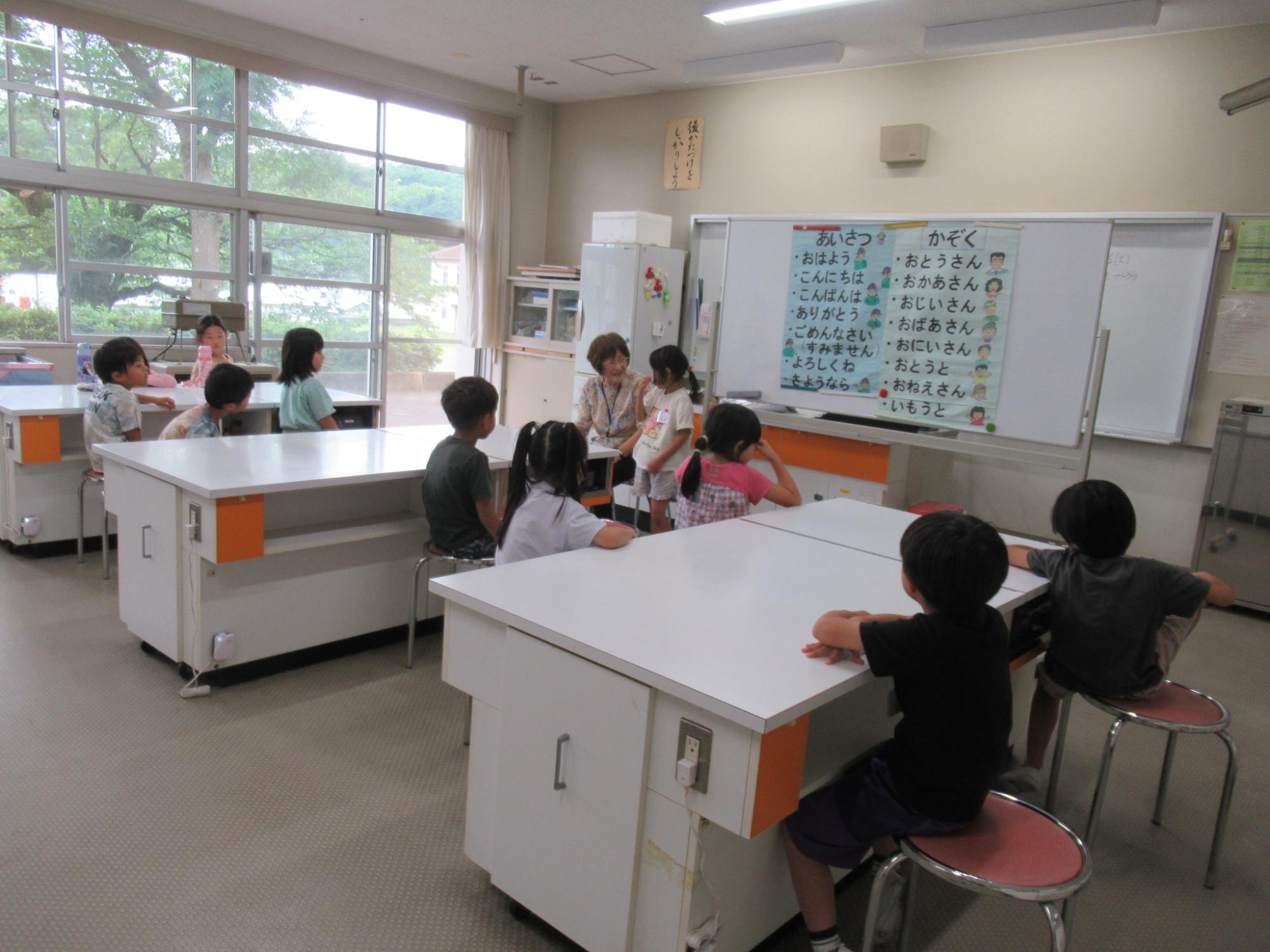 白とオレンジの実験台のような机が並ぶ教室で、ホワイトボードに挨拶と家族の日本語の単語リストが表示される中、数人の児童が教師を囲んで話を聞いている様子の写真