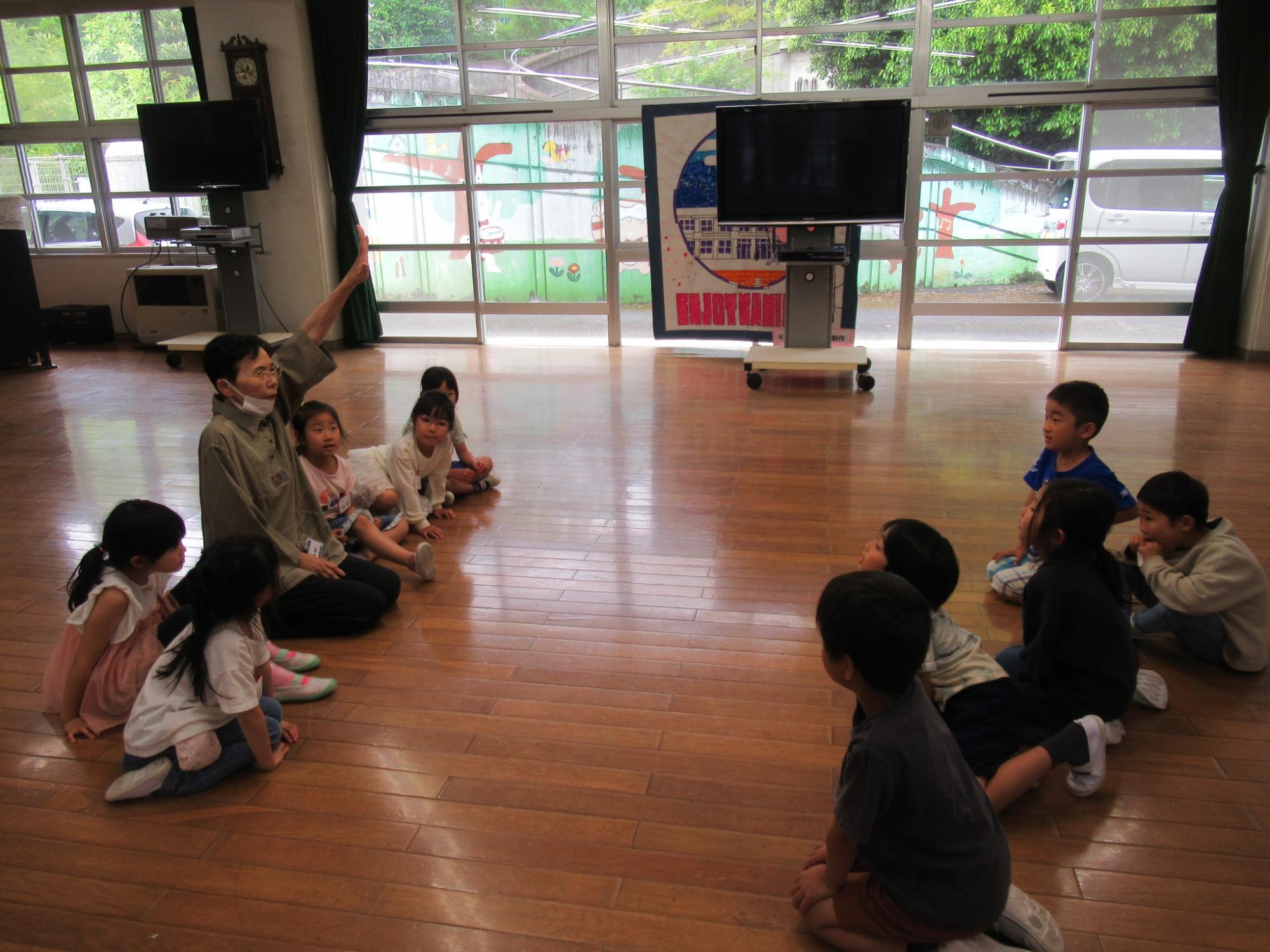 窓から明るい光が差し込むフローリングの広い室内で、中央に座った女性が右手を挙げて話し、その周りに床に座った約10人の未就学児らしき子どもたちが熱心に聞き入っている読み聞かせ会の様子の写真