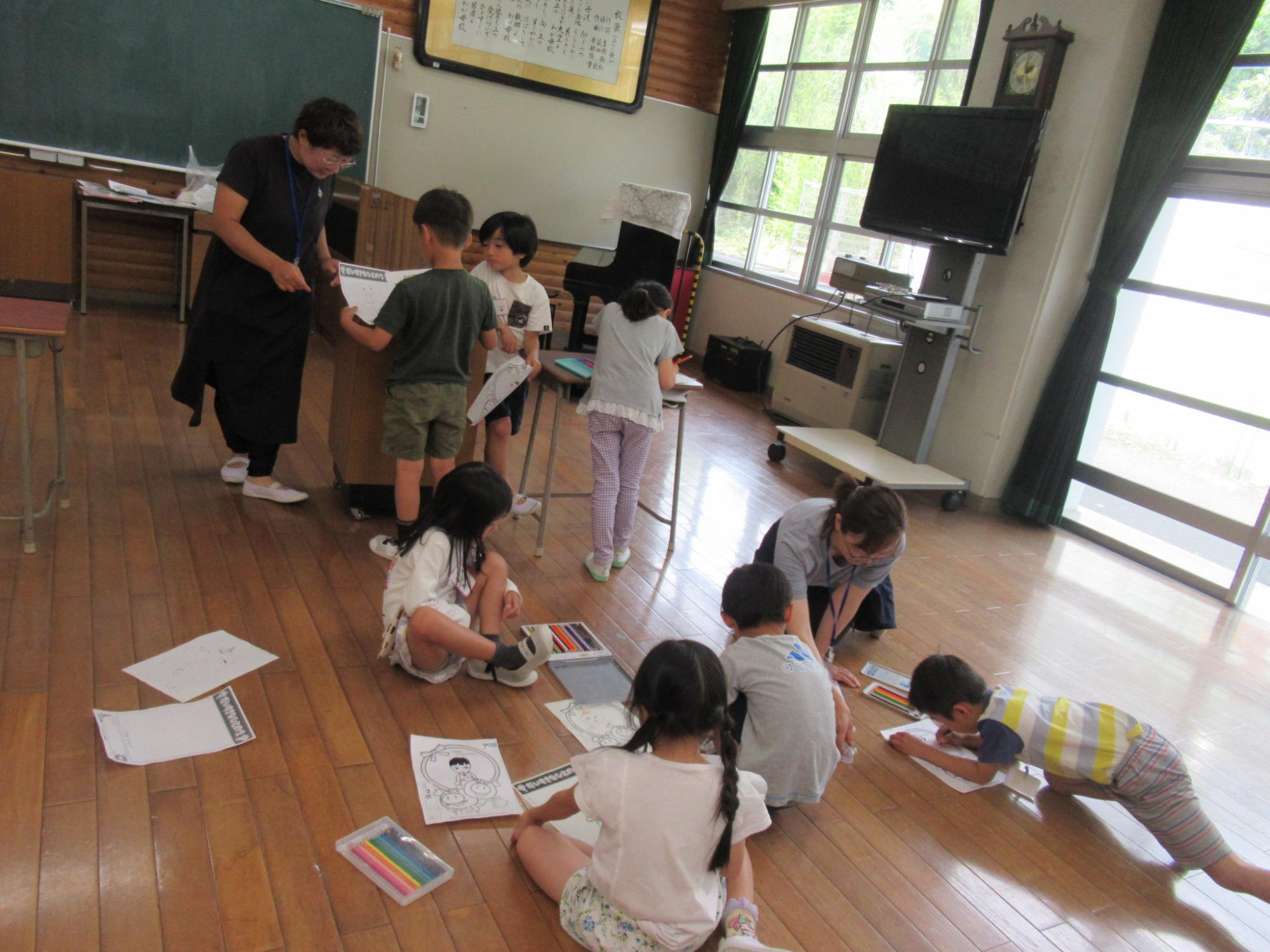 幼稚園のようなフローリングの広い室内で、黒い服を着た先生が立って子どもたちにプリントを渡しており、他の子どもたちはクレヨンや色鉛筆を使って床で絵を描いたり作業したりしている風景を捉えた写真