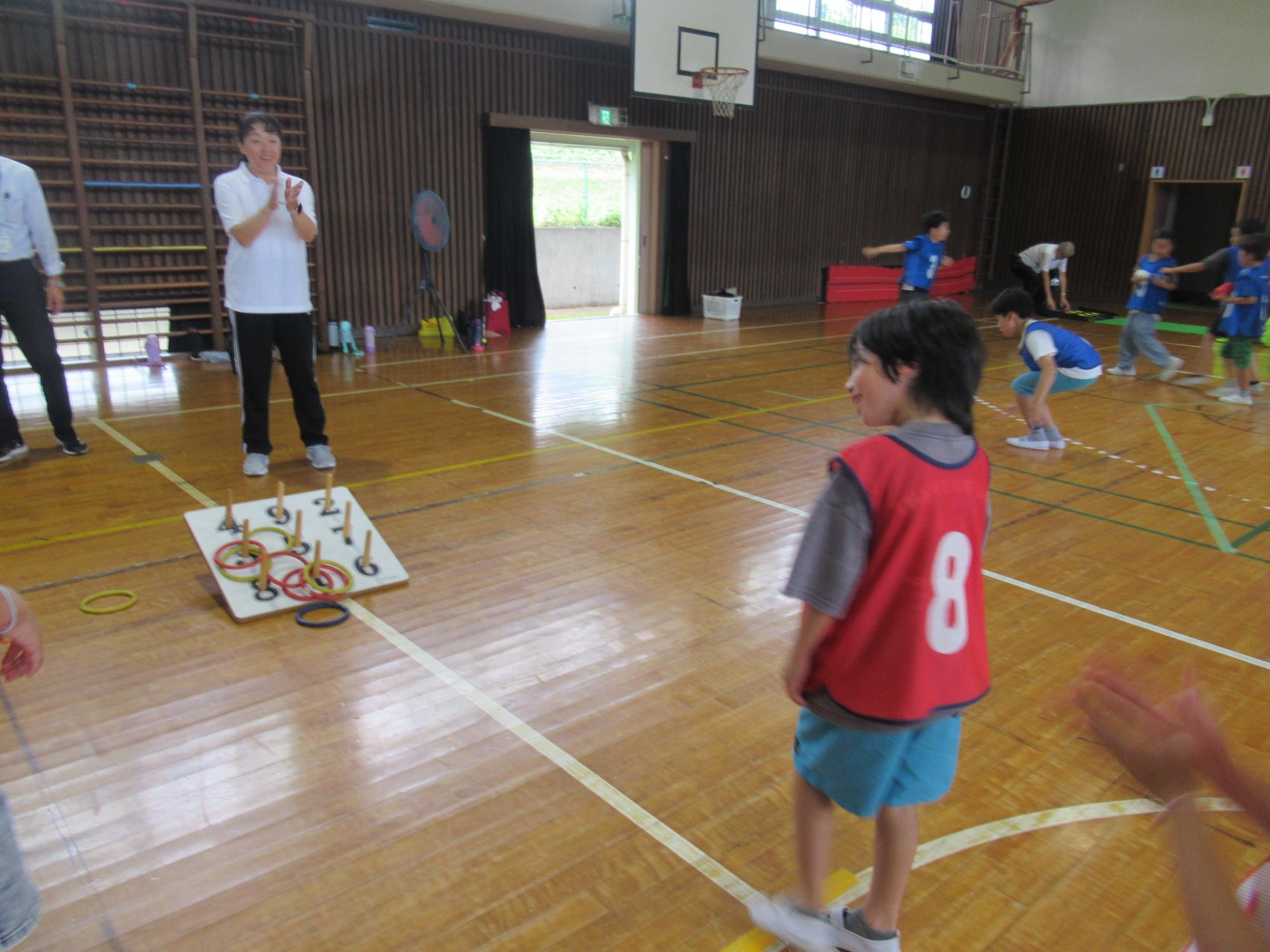 体育館の光沢のある木目調の床で、白いシャツを着た大人が拍手をしている傍らで輪投げの用具が置かれ、赤いビブスを着た男の子が振り返って笑っており、奥では青いビブスを着た子どもたちが走り回るなど、小学校の体育館でのレクリエーション活動の様子の写真