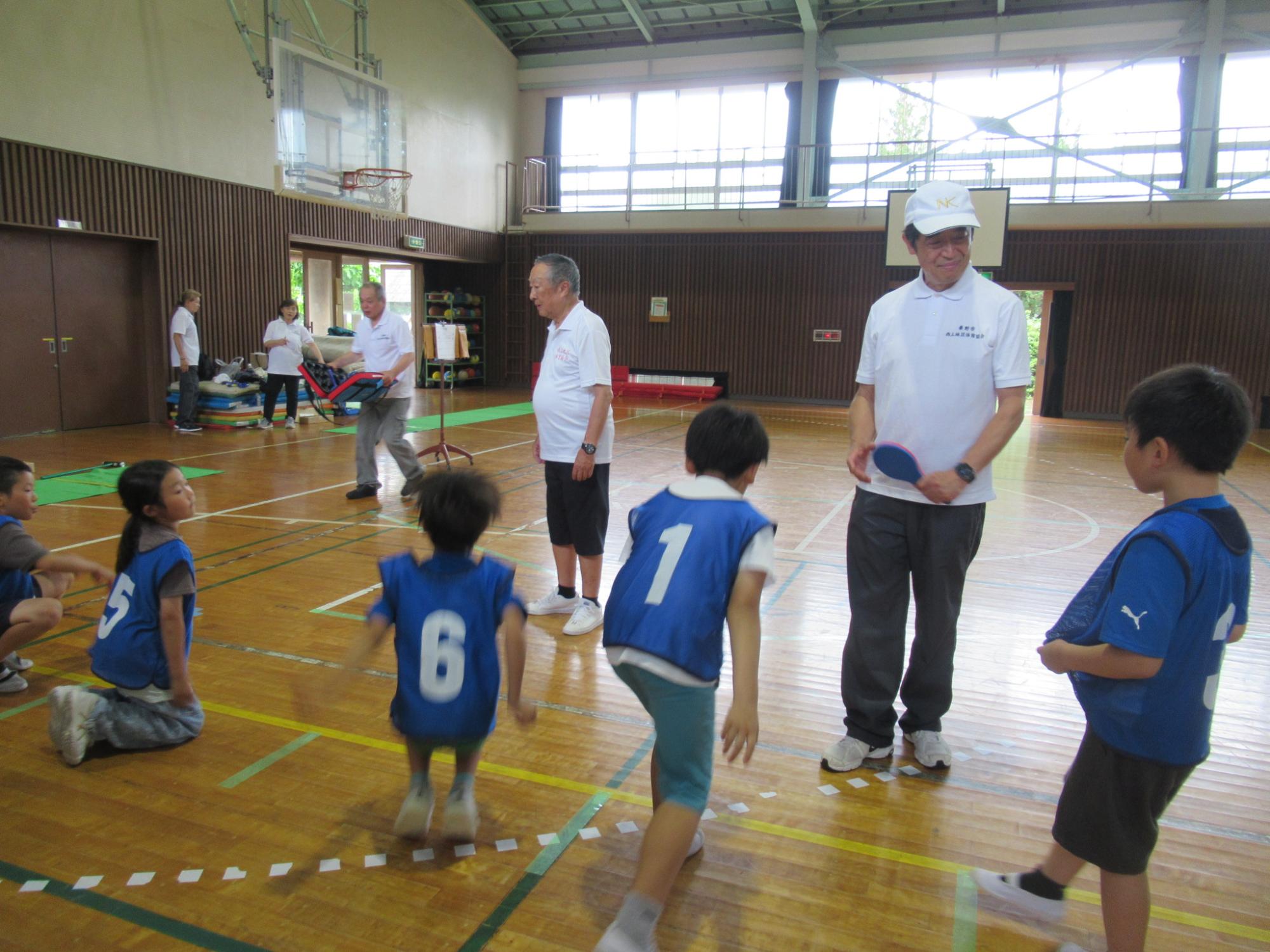 体育館の広い木目調のフロアで、青いビブスを着た数人の小学生ほどの子供たちが運動しており、右側には白いポロシャツと帽子をかぶった男性がラケットを持ち、高齢者と子どもたちが一緒に運動を楽しんでいるスポーツ交流会の様子の写真