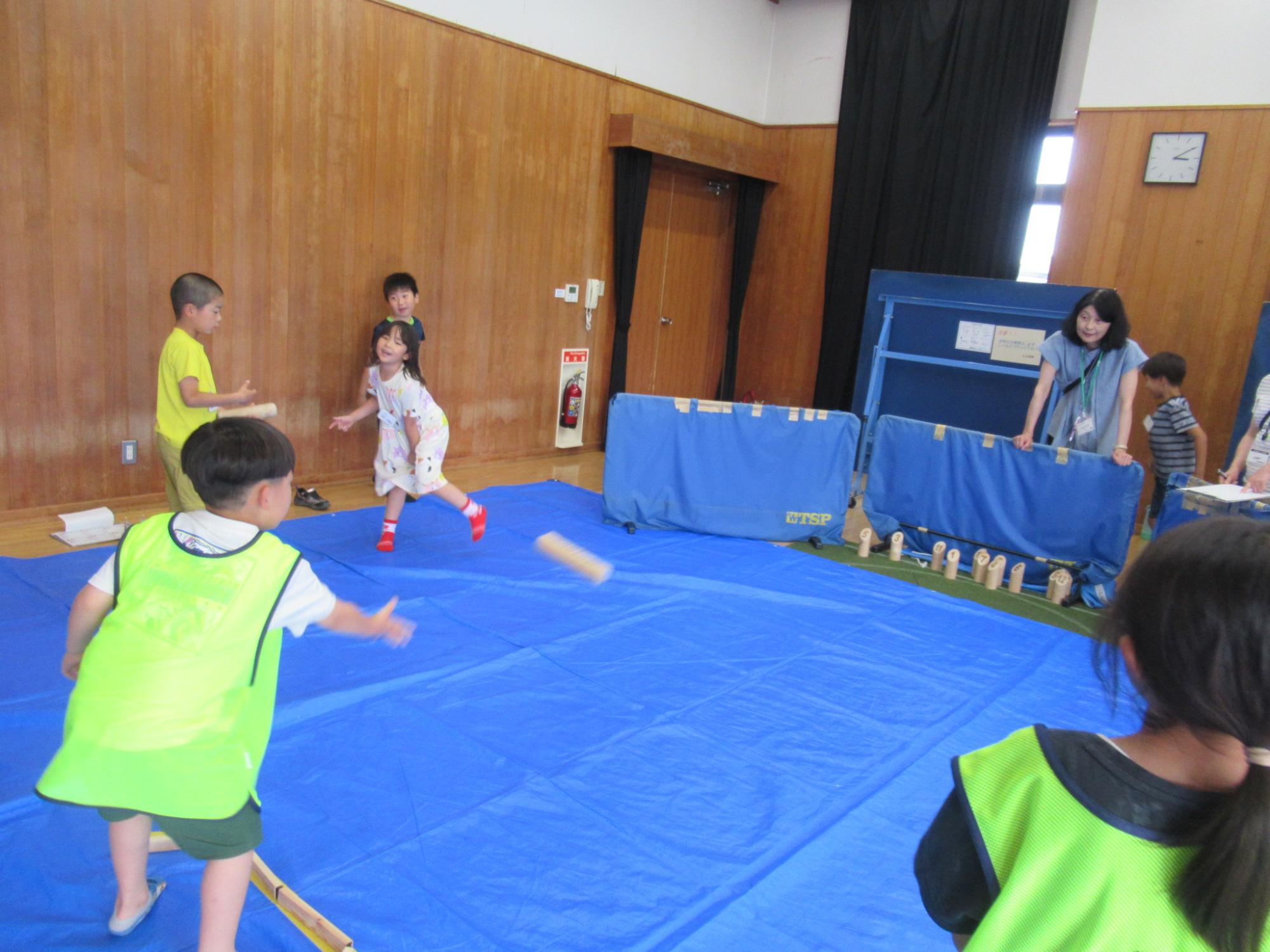 体育館のような木目調の壁の室内で、床に広げられた青いシートの上で、黄色いTシャツや蛍光黄緑色のビブスを着た小学校低学年ほどの子どもたちが、木片を投げて遊ぶゲームを楽しんでおり、後ろでは女性がそれを見守っているレクリエーション活動の様子の写真