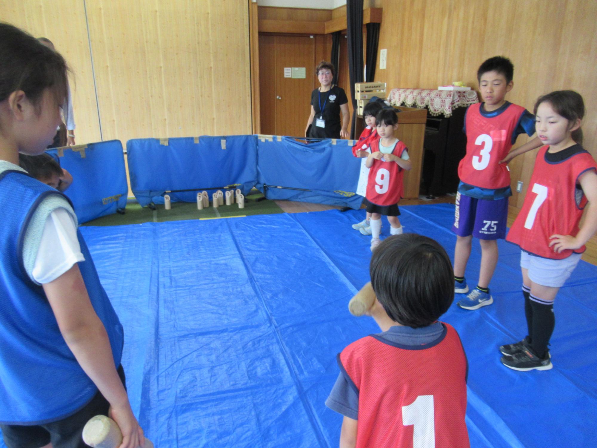 木目調の壁の室内で、床に敷かれた青いシートの上で、後ろに立つ女性が見守る中、赤いビブスと青いビブスを着た小学校低学年ほどの子どもたちが、木製の棒を持ってターゲットに向かって投げ、キャッチボールゲームのようなアクティビティを楽しんでいる活動風景の写真