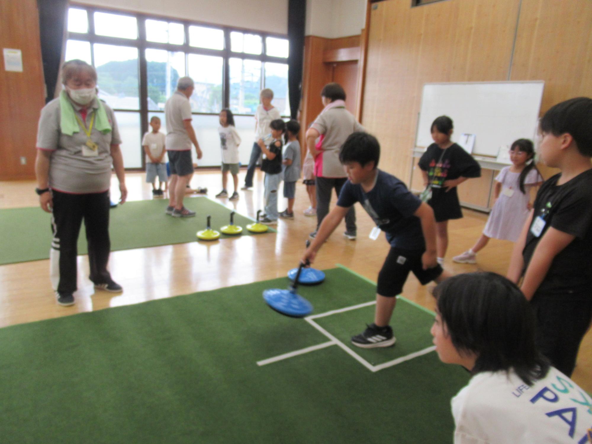 体育館のような広い室内で、床に敷かれた緑色のマットの上で、小学生ほどの子どもたちが青いディスクをスティックで押し出して投げる、カーリングのようなニュースポーツを楽しんでおり、周りでは大人たちが見守っている活動風景の写真